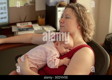 Mother works at a home office desk surrounded by computer and books while baby sleeps peacefully on her chest, work life balance Stock Photo