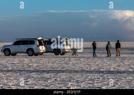Tour guides set up a folding champagne table beside two parked SUVs while travelers watch the sunset on the Salar de Uyuni salt flat, Uyuni, Bolivia Stock Photo