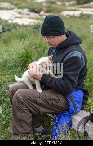Man in a black beanie holds a fluffy white Greenland Dog puppy gently on his lap on arctic tundra near Ilulissat, Greenland Stock Photo