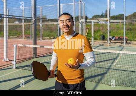 Asian man smiling on outdoor pickleball court, holding paddle and perforated yellow ball, ready to play the popular racket sport in sunny, active leis Stock Photo