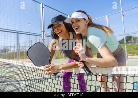 Two cheerful female friends standing by the net on a pickleball court, smiling and holding paddles and an orange ball after playing, symbolizing frien Stock Photo