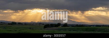 View of dramatic sun rays breaking through dark storm clouds over a mountain range and green fields at sunset United States. Stock Photo