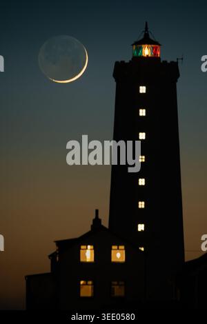 View of Grotta Lighthouse silhouetted against a twilight sky with a crescent moon and glowing windows in Seltjarnarnes, Seltjarnarnesbaer, Iceland. Stock Photo