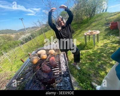 Woman joyfully dancing while cooking various meats and vegetables on an outdoor charcoal barbecue grill, celebrating a spring gathering surrounded by Stock Photo