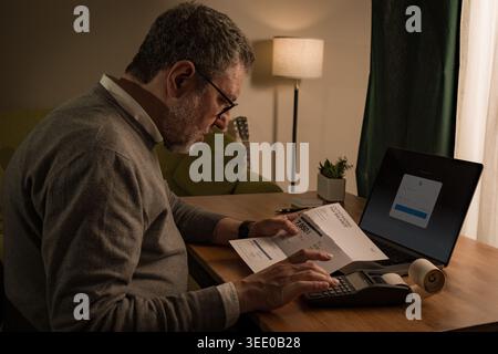 Senior man at home at night calculating bills and expenses with calculator and laptop, managing finances and reviewing documents under warm light. Stock Photo