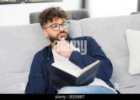 Thoughtful man in glasses and beard sitting on a comfortable sofa at home, reading a book and contemplating Stock Photo