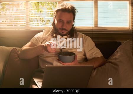 Young adult Caucasian man eating cereal while using laptop on sofa, combining home routine with screen time. Suitable for remote lifestyle, streaming, digital leisure marketing Stock Photo
