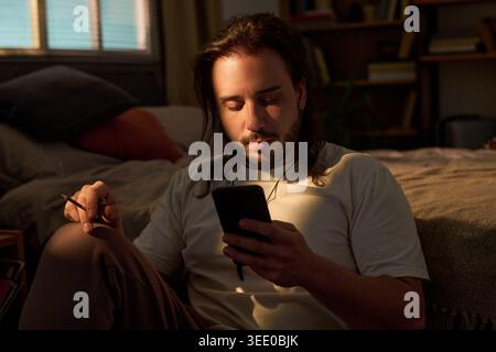 Young adult Caucasian man sitting on floor, checking smartphone while holding cigarette in bedroom, suggesting screen addiction or unhealthy habit. Useful for wellness or addiction campaigns Stock Photo