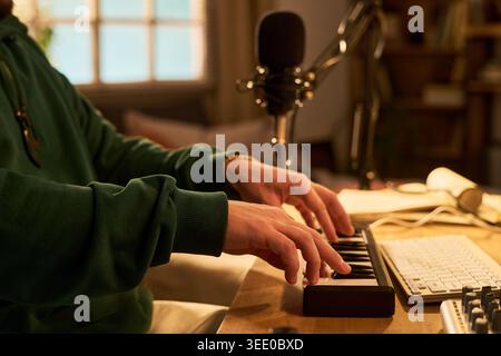Young adult man composing music on MIDI keyboard beside microphone and computer workstation, creating audio content. Ideal for podcasting, home studio, music production marketing Stock Photo