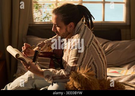 Young adult man writing lyrics in notebook while holding acoustic guitar on bed, side view. Conceptualizing songwriting and creative work for music education or artist branding Stock Photo