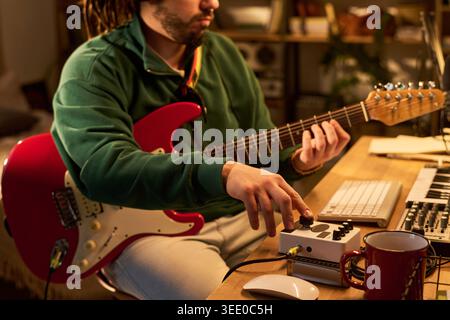 Young adult man playing electric guitar and adjusting audio interface in home studio, recording music content. Useful for music production, creator economy, audio technology marketing Stock Photo