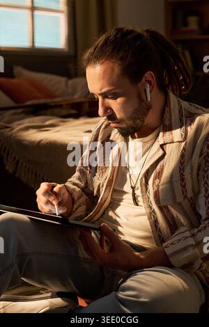 Vertical shot of young adult man using tablet while wearing wireless earbuds at home, browsing digital content. Suitable for remote lifestyle, streaming service, or mobile app marketing Stock Photo