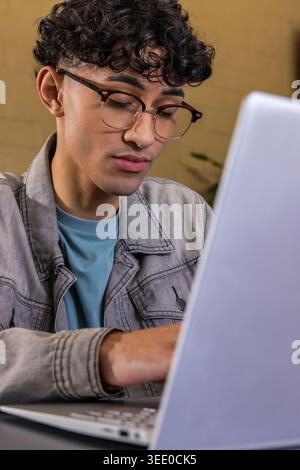 Teenage male working on silver laptop at desk in office, wearing denim jacket and teal shirt Stock Photo