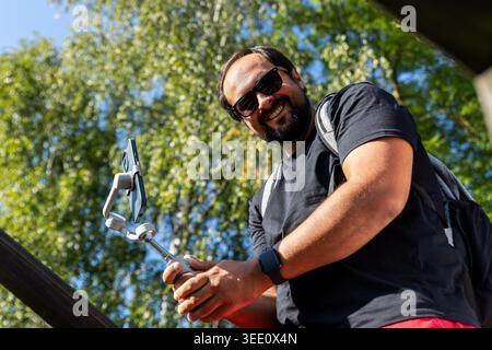 Man uses smartphone gimbal stabilizer filming outdoor video content sunny park green trees. Smiling vlogger create digital media modern equipment Stock Photo