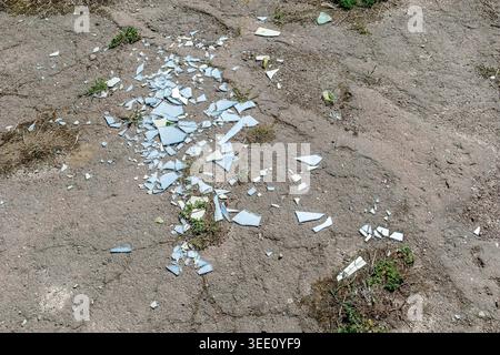 Scattered shards of broken glass lying on cracked asphalt ground with small patches of green grass. Stock Photo