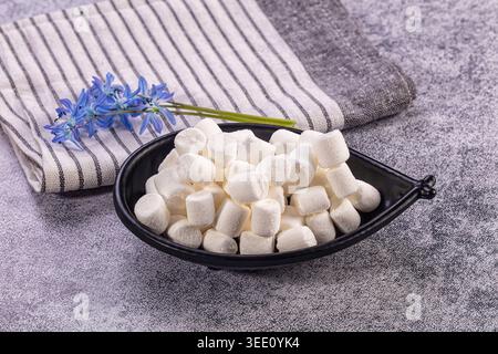 Fluffy white mini marshmallows fill black bowl. Striped grey, white napkin, delicate blue spring flowers rest beside Stock Photo