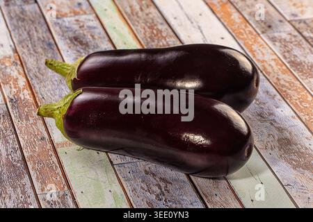 Two fresh purple eggplants displayed on rustic wooden surface. Top down composition highlights glossy deep violet skin vibrant green stems. Soft studi Stock Photo