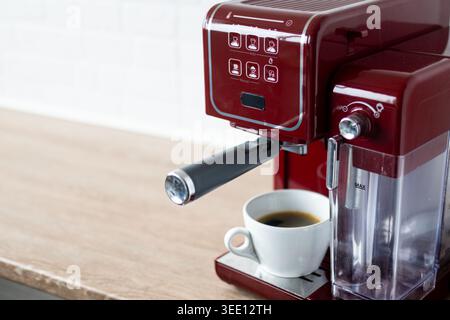 Close-up of a red espresso coffee machine brewing fresh coffee into a white cup on a wooden countertop in a modern kitchen. Minimal home coffee prepar Stock Photo