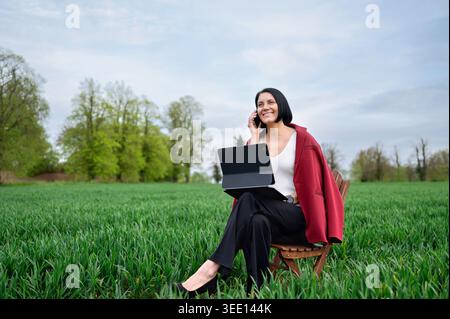 Woman using laptop and talking on smartphone in green field outdoors Stock Photo