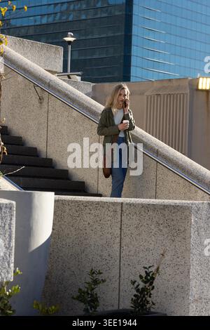 Woman descending concrete stairs at urban plaza holding coffee cup and smartphone, copy space. Pedestrian, city, daylight, glass, highrise, modern, architecture Stock Photo