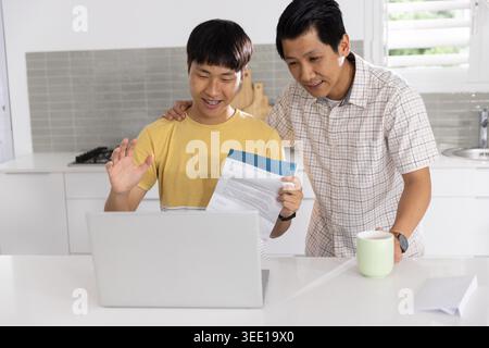 Asian family members reviewing documents on laptop at home kitchen with green mug and envelope. Duo, computer, papers, letter, cup, stock, interiors Stock Photo