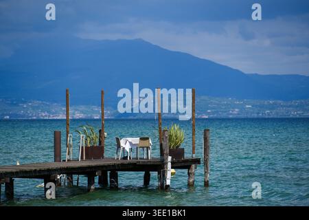 View of a table for two set on a wooden pier overlooking the blue waters of Lake Garda with mountains in the background Sirmione, Lombardy, Italy. Stock Photo