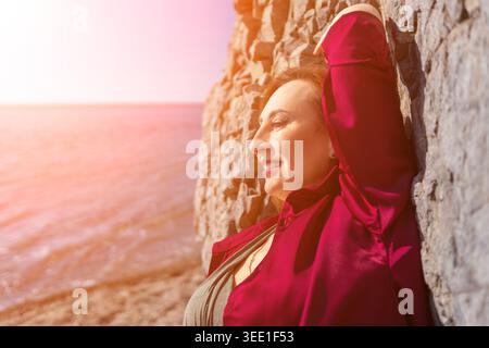 Woman smiling coastline mature lady in burgundy shirt leans against a stone wall on a sunny beach during sunset Stock Photo