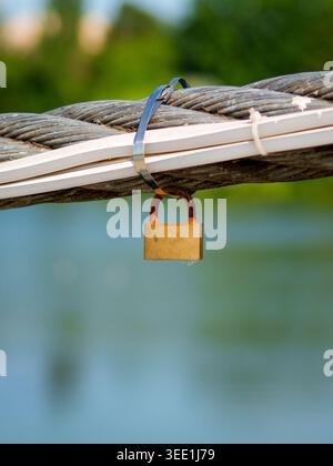 A small brass padlock with visible rust on its shackle hangs from a thick braided steel cable secured with a metal zip tie. Stock Photo