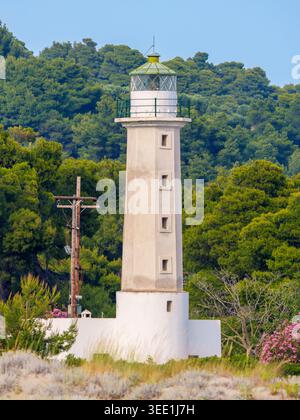 A white stone lighthouse with a green-topped lantern room stands against a densely forested hillside under a clear blue sky. Stock Photo