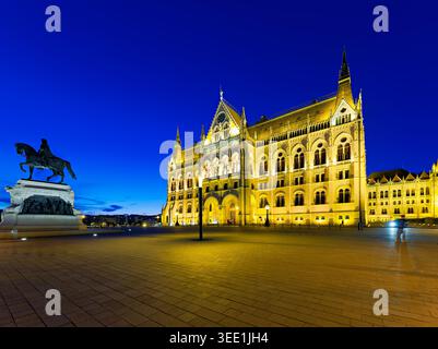 Budapest Parliament building stands tall at night with lights shining bright. A statue is visible in the foreground as the sky turns dark. Stock Photo