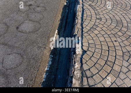 Urban street showing contrast between rough asphalt road and structured cobblestone pavement separated by dark gutter trench evoking stark rugged and Stock Photo