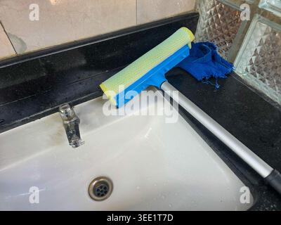 Blue squeegee with yellow sponge resting on white ceramic sink for daily cleaning chore in modern bathroom promoting good hygiene and fresh domestic e Stock Photo