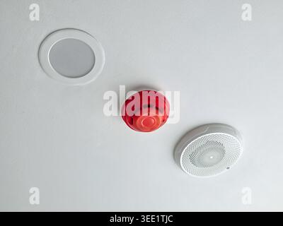 Red fire alarm smoke detector mounted on white ceiling next to lighting and sensor providing calm secure feeling of indoor safety and building protect Stock Photo