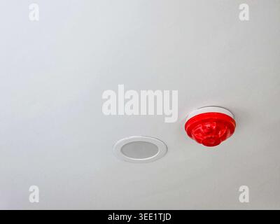 Red fire alarm mounted on white ceiling providing safety sensor and smoke detector function giving secure feeling alongside audio equipment in buildin Stock Photo