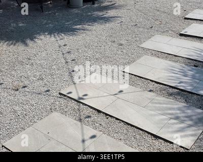 Rectangular concrete stepping stones arranged on gravel ground creating pathway with shadows of string lights overhead giving serene and peaceful outd Stock Photo
