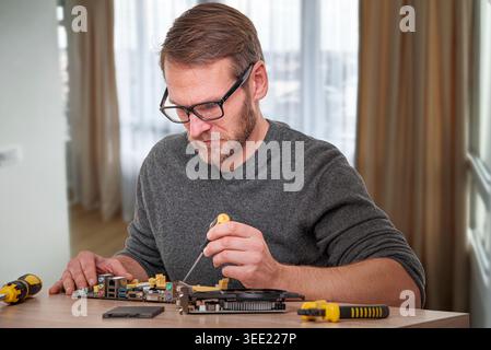 A focused technician with glasses uses a screwdriver to repair a computer motherboard and graphic card on a desk. Stock Photo