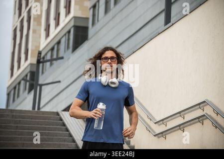 Athletic man with water bottle standing near stairs after outdoor training session in modern city environment Stock Photo