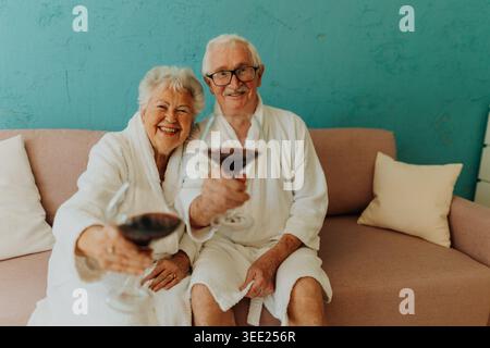 Happy senior couple sitting together in bathrobe on sofa with glass of wine. Stock Photo