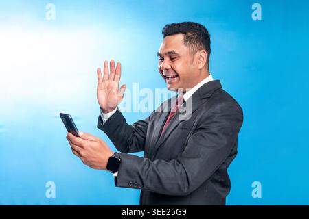 Friendly Indonesian executive in a suit waving his hand while having a video call on a smartphone against a blue studio background. Stock Photo