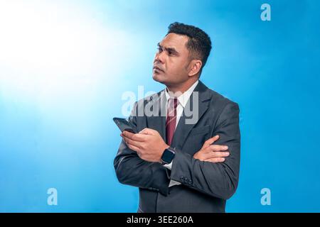 Portrait of an Indonesian executive in a suit, holding a smartphone with a skeptical and pensive expression against a blue studio background. Stock Photo