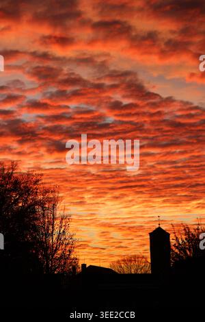 Silhouetted barn stands against fiery, cloud-filled sky. Rays of orange glow paint the horizon. Trees frame the scene in dark, leafless branches. Mood Stock Photo