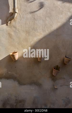 Rustic textured wall with hanging terracotta pots and dramatic diagonal shadows in a historic Mediterranean village house exterior under bright sunlig Stock Photo
