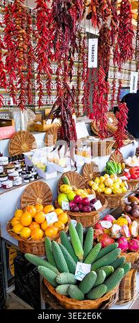 Vibrant red chiles hang in rows above the stall. Fresh cucumbers, lemons, and oranges sit in woven baskets. Vendor stands nearby, offering produce to Stock Photo