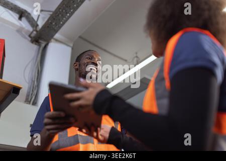 Two African American factory workers discuss production workflow on a tablet while working with industrial machinery at manufacturing plant. Stock Photo