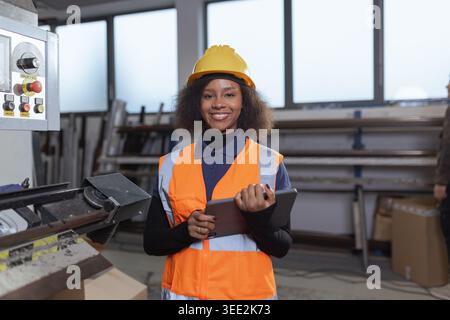 African American woman portrait next to machinery and digital tablet use in a factory setting while ensuring production quality. Stock Photo