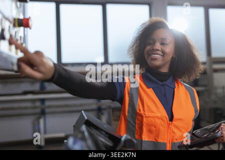 African American woman working with machinery and uses a digital tablet in a factory setting. She focuses on quality control and safety. Stock Photo