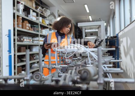 A female factory worker working on machinery while using a digital tablet in a modern industrial setting. Stock Photo