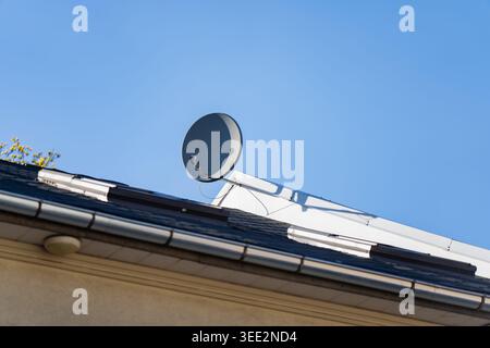 Black satellite dish antenna mounted on the dark shingle roof of a residential house for telecommunications and digital broadcasting reception against Stock Photo