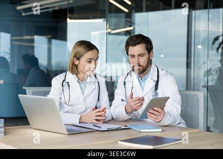 Two doctors wearing stethoscopes and lab coats collaborating on a research project, discussing health data and patient records displayed on a digital tablet in a modern office Stock Photo