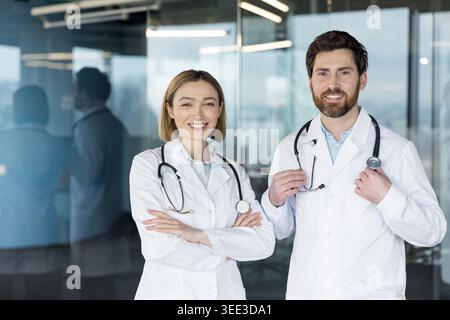 Two medical staff members, a man and a woman in white lab coats with stethoscopes, standing together smiling, representing trust, healthcare, and professional support in a clinical setting Stock Photo